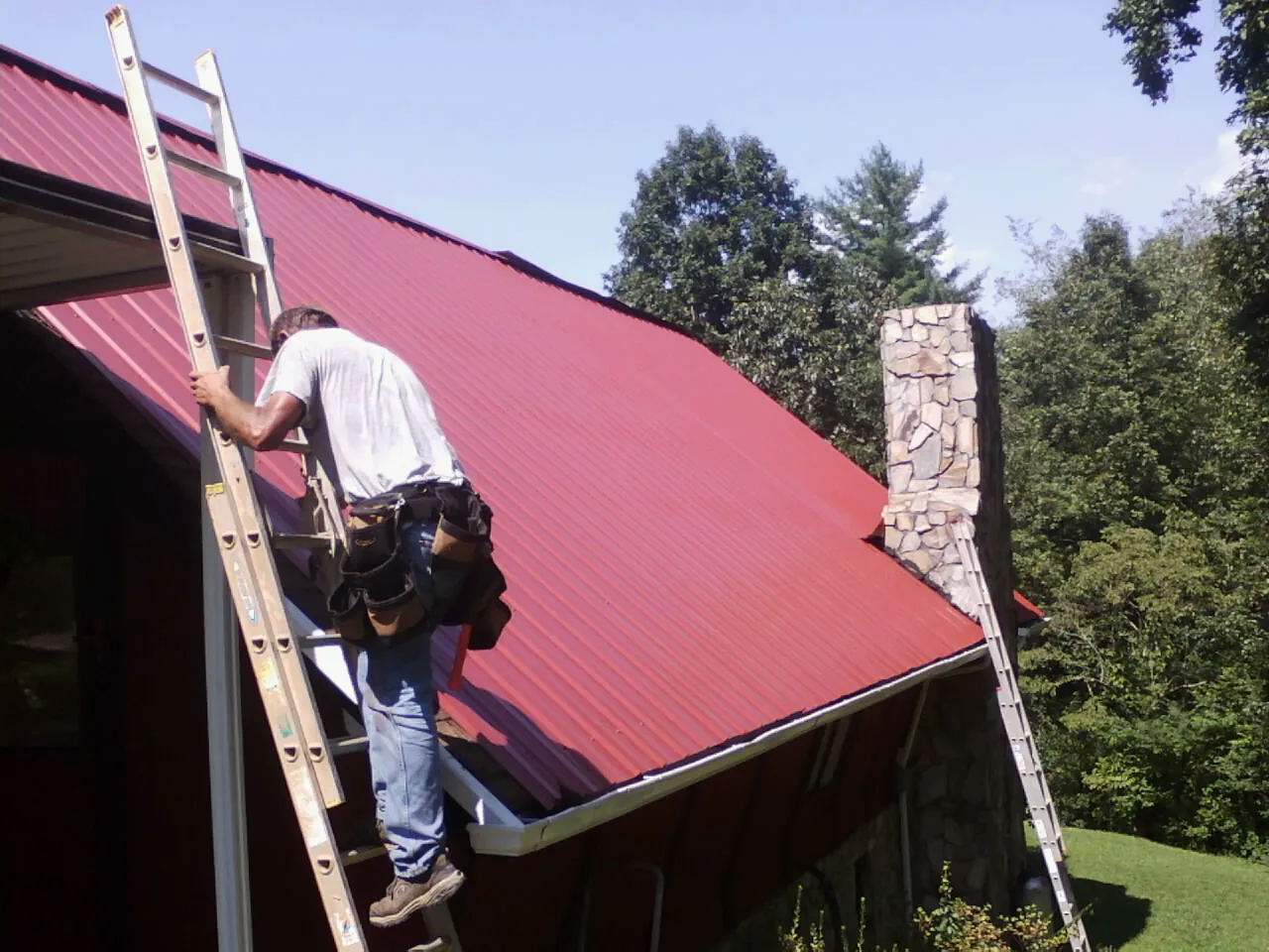 Roofer on a ladder beside a red metal roof and stone chimney — Soffit & Fascia Repair by Roofing Brothers in Wasco